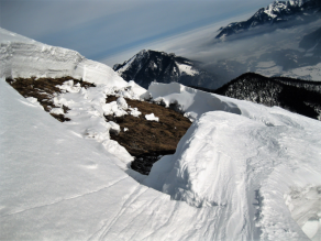 Die mächtige Schneedecke öffnet sich zu einem Gleitschneemaul. Der grasige Boden wird sichtbar. Im Hintergrund sieht man das winterliche Chiemgau mit Nebelresten im Tal.