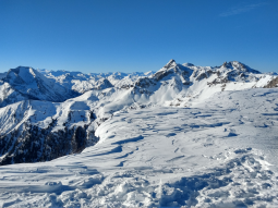 Wind hat den Schnee im Vordergrund stark bearbeitet. Es haben sich Windgangeln gebildet. Im Hintergrund sind die Gipfel der Berchtesgadener Alpen vor wolkenlosem Himmel zu sehen.