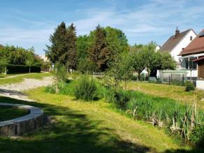 Naturnah verlaufender Bach mit Hochstauden-, Gras- und Gehölzvegetation am Ufer. An einer Seite grenzen Gärten und Wohnbebauung an, auf der anderen Seite parkartiges Gelände mit einer Sitzgelegenheit aus quaderförmigen, halbkreisförmig angeordneten Natursteinen und ein neu gepflanzter Baum.