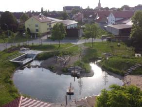 Neu gestalteter Teich mit Wassertretanlage, Spielmöglichkeiten mit Floß und Sandstrand. Im Hintergrund Häuser und Straßen eines Ortes.