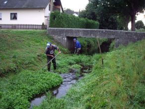 Graben, dessen Bewuchs auf der Sohle von zwei Männern mit einer Sense in der Hand entfernt wird.