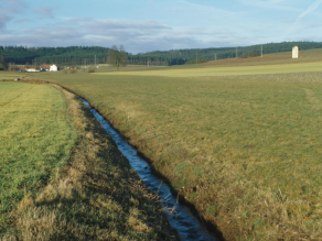 Stark begradigten Bach ohne Ufergehölze oder Uferrandstreifen in einer landwirtschaftlich intensiv genutzten Landschaft.