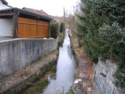 Bach mit hart verbauten Ufern (verfugte Steine); Betonmauer und Gebäude direkt am Ufer im linken Bildbereiche; Steinmauer und hohe Fichten direkt am Ufer.