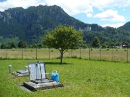 Messschacht eines Brunnens der Wasserversorgung neben dem eine Probenbox steht. Im Hintergrund Schloss Neuschwanstein.
