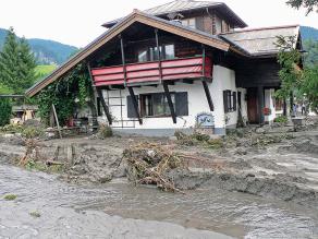 Zerstörte Häuser/Straße nach Abfluss eines Hochwassers. Neben den Schäden ist auch die Ablagerung von vom Hochwasser mitgeführtem Material zu sehen.