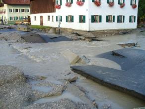 Hochwasserbedingte Schäden an einer Straße. Der Asphalt wurde teilweise unterspült, sodass keine ebene Fläche mehr vorhanden ist. Tiefe Senken und Löcher haben sich gebildet, durch die das Wasser abfließt.