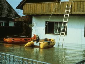Bei einem Hochwasserereignis stehen zwei Schlauchboote zur Evakuierung eines Wohnhauses im überschwemmten Vorgarten bereit. Über eine Leiter, die zum Balkon führt können die Bewohner das Haus verlassen und in die Boote steigen