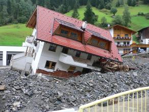 Haus in Vorarlberg (Österreich), das durch ein Hochwasser in Schräglage geraten ist und weggeschwemmt wurde. Das Haus ist umgeben von Geröll und größeren Felsbrocken, die durch das Hochwasser mobilisiert wurden.
