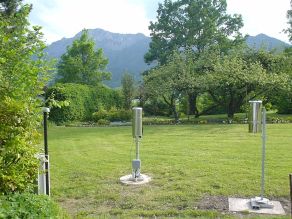 In einer Gartenlandschaft steht ein Schaltschrank mit Funkantenne, ein automatischer Niederschlagsmesser und ein Niederschlagsmesser für die manuelle Kontrollmessung
