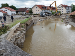 Ein Gewässer, bei dem die Ufermauern bzw. Uferbefestigungen zu großen Teilen eingestürzt sind. Etwa in der Mitte, wo vermutlich einmal ein Deich gestanden ist, klafft eine Lücke. Ein Bagger lädt Material auf einen bereit stehenden Lastwagen