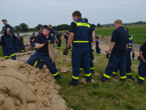 Es sind mehrere Personen (Einsatzkräfte) zu sehen, die Sandsäcke auf einem Deich aufschichten.