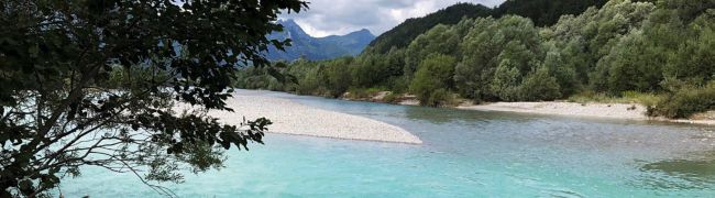 Ein Gebirgsfluss mit stark variierenden Wassertiefen, erkennbar an den unterschiedlichen Farben des Wassers (türkis, hellblau, dunkelblau...). In der Mitte befindet sich eine große Kiesbank. Die Ufer sind flach und strukturreich, dahinter schließt sich Auwald an