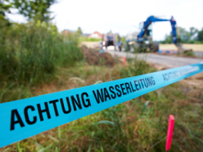 Absperrband mit Hinweis 'Achtung Wasserleitung' vor einer Baustelle mit Bagger.