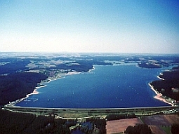 Die Luftaufnahme zeigt den Brombachsee mit seinen zwei Vorsperren - Kleiner Brombachsee und Igelsbachsee - im Hintergrund des Bildes. Das Absperrbauwerk erstreckt sich über die ganze Breite des unteren Bildrands.