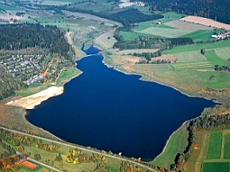 Das Luftbild zeigt den Perlsee inmitten der von Grünland geprägten Landschaft des Oberpfälzer Waldes; im Vordergrund das Absperrbauwerk und am linken, oberen Bildrand ein Campingplatz.
