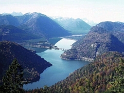 In der Luftaufnahme sieht man im Vordergrund den fjordartigen Sylvensteinspeicher dahinter die Berge des Karwendelgebirges. Das Sperrenbauwerk befindet sich am unteren, linken Bildrand.