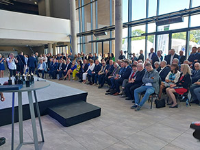 Blick auf das Foyer der Messe, ca. 100 Ehrengäste sitzen bzw. stehen in insgesamt vier Reihen vor der Empfangsbühne 