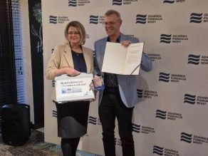 Ehrung des Vertreters des TTW; Leiterin der Wasserstiftung Danzig (GFW) mit dem Kollegen des LfU vor einem Banner der GFW mit Übergabe der Auszeichnung. Einer Urkunde und eines Glaskubus.