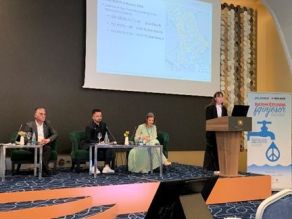 Referenten und Moderator des Panel Gewässerschutz/ Klärschlammmanagement auf der Bühne des Tagungsraumes, die meisten auf Stühlen sitzend, Frau Gega/ UNIBW am Rednerpult beim Vortrag.