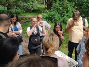 Teilnehmer der Sommerakademie im Kreis stehend mit der Referentin des WWA Hof mit Blick auf ein Becherglas bzw. Wannen, in denen sich Sediment sowie Kleinlebewesen des Baches befinden.