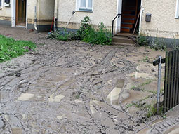 Ablagerung von Schlamm in und an einem Wohnhaus mit deutlich sichtbarer Anschlagslinie, die den höchsten Hochwasserstand auf ca. 60 cm über Geländeoberfläche zeigt.