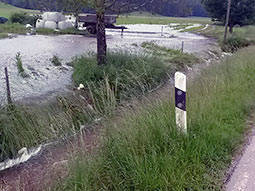 Wild abfließendes Wasser von einer Wiese strömt nach einem Starkregenereignis auf der Geländeoberfläche in Richtung eines Straßengrabens und füllt diesen bordvoll.